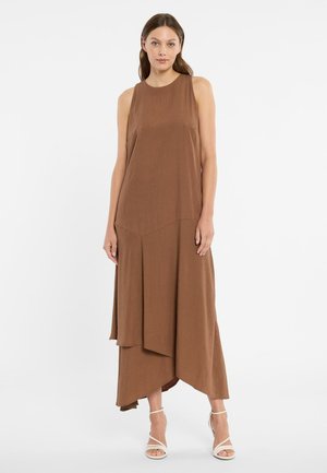 Frau trägt ein ärmelloses, braunes, asymmetrisches Midikleid und weiße, Riemchen-High-Heel-Sandalen, steht vor einem einfarbigen weißen Hintergrund.