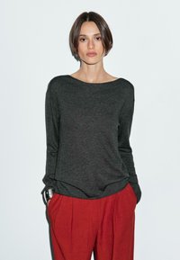 Une femme aux cheveux bruns courts se tient devant un fond clair, portant un pull sombre et un pantalon rouge, les mains dans les poches.