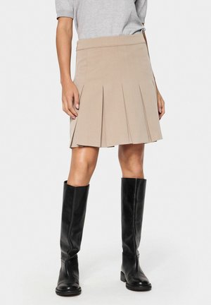 Beige pleated skirt with a fitted waist, made of textured fabric. Paired with knee-high black leather boots.
