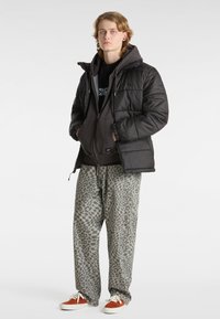 Schwarze Pufferjacke, über einem dunklen Hoodie getragen. Lockere, gemusterte graue Hose mit Leopardenmuster. Turnschuhe mit orangefarbenen Akzenten.