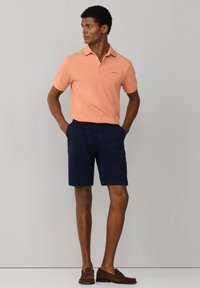 Homme debout les mains dans les poches, portant un polo saumon, un short marine et des mocassins marron, sur un fond uni.