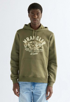 Junger Mann trägt einen olivgrünen Wrangler-Hoodie mit Vintage-Auto-Grafik und hellblauen Jeans, steht vor einem schlichten weißen Hintergrund.