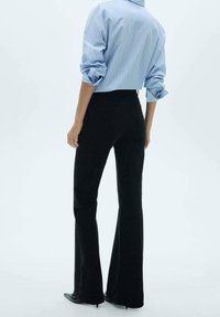 Zwarte geplooide broek van gladde stof, gecombineerd met een lichtblauw gestreept overhemd met knopen. De mouwen van het overhemd zijn opgerold.