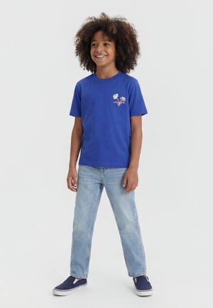 Smilende barn med krøllet hår iført en blå t-shirt med grafik, lyseblå jeans og marineblå slip-on sko stående mod en ensfarvet baggrund.