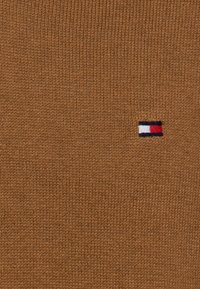 Brauner gestrickter Pullover mit einem kleinen gestickten Logo und roten, weißen und blauen Streifen auf der linken Seite. Weicher Stoff und engmaschiges Design.