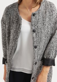 Cardigan de tweed gris y blanco con un tejido texturizado, cierres de botón negros y puños negros en contraste. Llevado sobre una camiseta blanca.