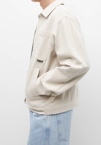 Beige bomuldjakke med knaplukning, brystlomme og sidelommer; har en afslappet pasform og synlige syninger.