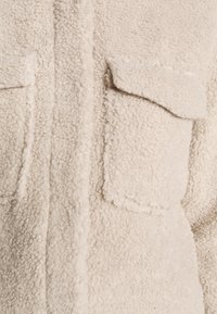 Zbliżenie beżowego materiału sherpa z kieszenią zakładkową oraz teksturowaną, miękką, kręconą powierzchnią.