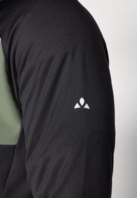 Schwarz langärmliges Shirt mit grünem Akzent auf der Schulter, ausgestattet mit einem strukturierten Stoff und einem kleinen weißen Logo am Ärmel.