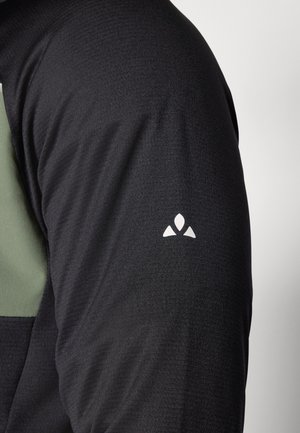 Chemise noire à manches longues avec un accent vert sur l'épaule, en tissu texturé et avec un petit logo blanc sur la manche.