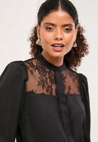 Mustast pluusist, milles on läbipaistev pitsipaneel lillemustritega, puff-särgid ja nööbitav kaelus. Pehme, tekstuurne kangas.