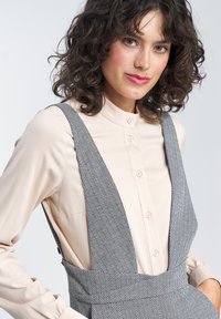 Szare ogrodniczki w jodełkę z szerokimi ramiączkami, noszone na jasnobeżowej, zapinanej koszuli. Gładka faktura i klasyczny krój.