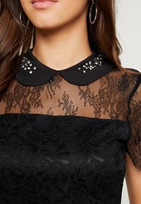Mujer vistiendo una blusa negra de encaje con un patrón floral transparente y un collar Peter Pan negro decorado, además de llevar pendientes de aro plateados.