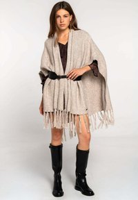 Poncho de punto beige con flecos en la parte inferior, asegurado con un cinturón negro en la cintura. Combinado con botas altas negras hasta la rodilla.