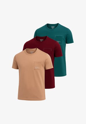 Tres camisetas de manga corta en color beige, burdeos y azul verdoso, hechas de algodón, con un bolsillo en el pecho que presenta texto. Textura de tela suave.