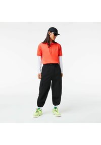 Červené polo tričko s krátkým rukávem, černé cargo kalhoty, zelené a bílé tenisky a černá čepice. Oblečení má logo Lacoste.