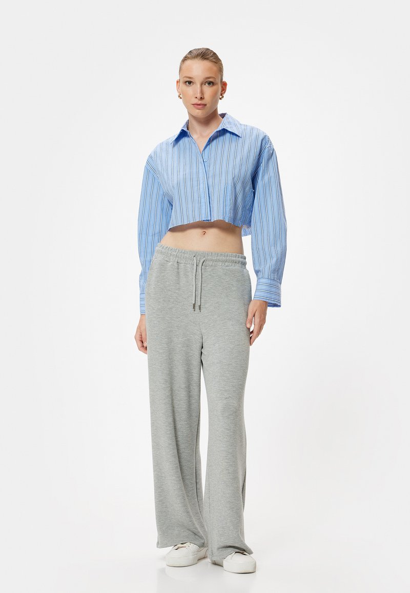 Gekürztes blau-weiß gestreiftes Hemd mit Knopfverschluss, kombiniert mit grauen Sweatpants. Das Model trägt weiße Sneakers. Einfaches, lässiges Design.