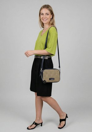 Lächelnde Frau in grünem Oberteil und schwarzem Rock geht und trägt schwarze Sandalen mit Absatz sowie eine gemusterte Umhängetasche mit schwarzem Riemen vor grauem Hintergrund.