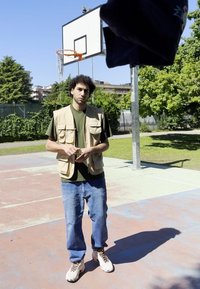 Béžová vesta s viacerými vreckami cez zelenú košeľu, modré džínsy a biele tenisky. V pozadí basketbalový kôš, slnečné vonkajšie prostredie.