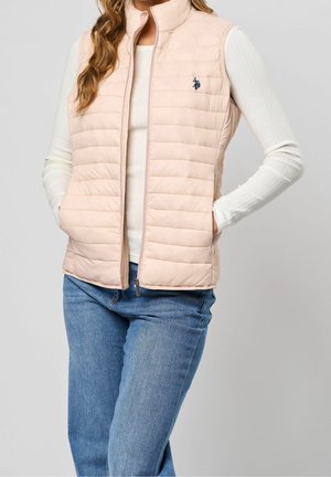 Femme portant un gilet matelassé rose clair sur un haut blanc à manches longues et un jean bleu, debout les mains dans les poches du gilet.