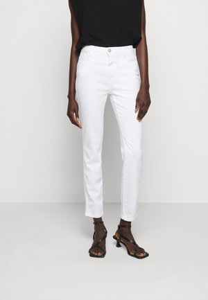 Femme portant un pantalon blanc ajusté longueur cheville, un haut noir sans manches et des sandales à talons noires à brides, debout devant un fond blanc uni.