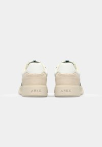 Białe i beżowe sneakersy z zamszowym obcasem, siateczkowymi bokami i gumową podeszwą. Posiadają minimalne oznakowanie i zaokrąglony kształt.