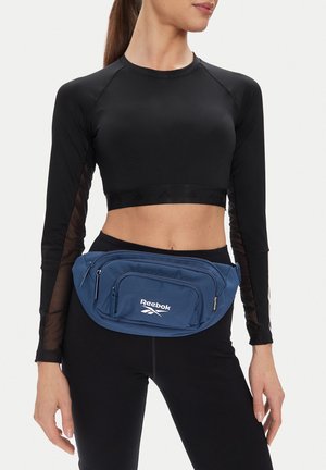 Kobieta ubrana w czarny krótki top i legginsy, z niebieską saszetką Reebok przypiętą wokół bioder.