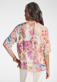 Mujer con cabello largo y gris que lleva una blusa de manga corta con estampado floral y abertura en forma de llave en la espalda, y pantalones blancos, de pie con la mano en la cadera.