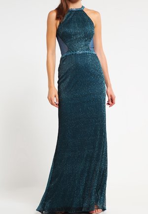 Bezrekawowa błyszcząca sukienka wieczorowa w kolorze zielono-niebieskim z dekoltem halter i przezroczystymi panelami po bokach, noszona przez stojącą osobę na białym tle.