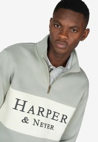 Hellgrüner Pullover mit weißem Querstreifen, auf dem der Text "HARPER & Neyer" steht. Mit Reißverschluss am Kragen und gestreiftem Shirt darunter.