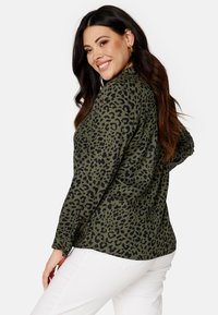 Camicia verde oliva con stampa leopardata nera, maniche lunghe e polsini con bottoni, abbinata a pantaloni bianchi. Tessuto morbido, vestibilità rilassata, senza distintivi particolari.