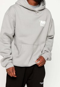 Grå huvtröja med ficka fram, med en tryckt vit logotyp på bröstet. Tillverkad av mjukt tyg med ribbade ärm- och nederkant.