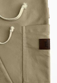 Beige stof met een diagonaal ribbelpatroon, een voork pocket en een donkerbruin leren label met reliëfdetail, inclusief een trekkoord.