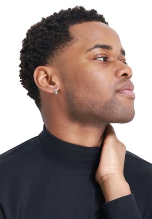 Boucle d'oreille en argent à l'oreille gauche, haut noir à col montant, peau lisse et cheveux courts et bouclés à la mode, vus de profil sur fond blanc.
