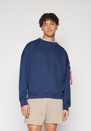 Junger Mann mit kurzen Dreadlocks, der ein marineblaues Sweatshirt mit langen Ärmeln und beige Shorts trägt, vor einem schlichten hellen Hintergrund stehend.