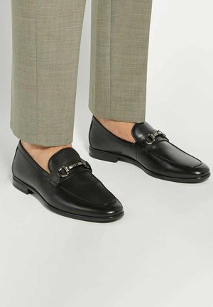 Black leather loafers with a flat sole, featuring a metallic buckle detail and rounded toe. Worn with light gray tailored pants.