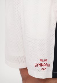 Balti tinkliniai sportiniai šortai su perforacijomis, su raudona siuvinėta etikete "MILANO GYMNASIUM EA7" ir juodu šoniniu paneliu.