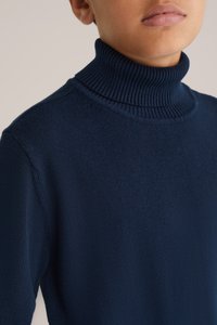 Temno modra turtleneck kapa iz mehke pletene tkanine. Ima rebrast ovratnik in gladko teksturo. Prikaz v bližnjem planu se osredotoča na zgornji del oblačila.