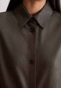 Brązowa skórzana koszula z wyprofilowanym kołnierzem, z zapięciem na guziki z przodu oraz czterema czarnymi guzikami. Gładka tekstura z widocznymi detalami przeszyć.