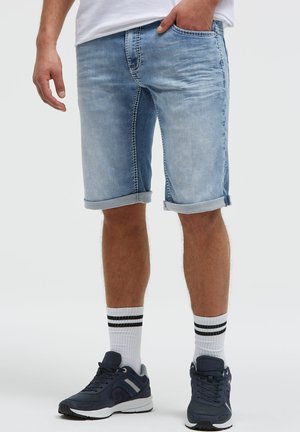 Mann trägt hellblaue hochgekrempelte Jeansshorts, weiße Socken mit schwarzen Streifen und dunkelblaue Sneakers, steht mit einer Hand in der Tasche.