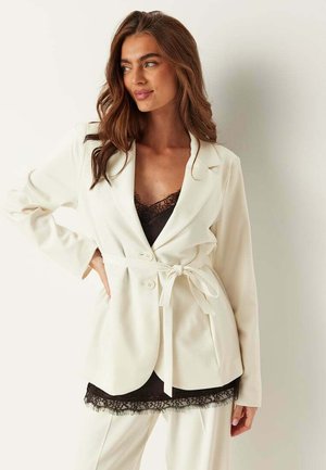 Kvinde iført en hvid blazer med bælte over en sort top med blondekant og matchende hvide bukser, stående med den ene hånd på hoften.