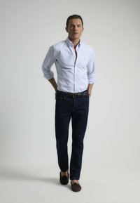 Camisa de botones a rayas azul claro, jeans azul oscuro, cinturón marrón y mocasines oscuros. El modelo se encuentra de pie con las manos en las caderas frente a un fondo claro.