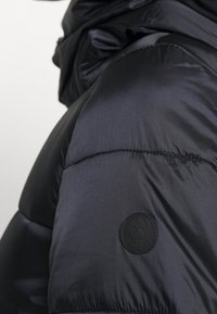 Schwarze Steppjacke mit glatter, glänzender Oberfläche, gesteppten Abschnitten und einem markanten runden Logo-Patch.
