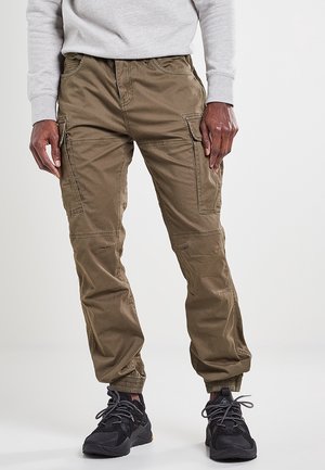 Mężczyzna ubrany w khaki spodnie typu cargo i czarne trampki stoi na jasnym, jednolitym tle.