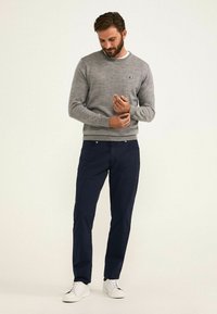 Suéter gris de punto con cuello redondo, combinado con pantalones chinos azul marino y zapatillas blancas, de pie frente a un fondo claro.