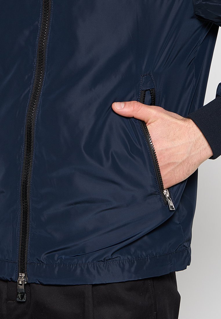 Marineblaues, leichtes Jacket mit einer glatten, glänzenden Textur, ausgestattet mit einem Frontreißverschluss und einer seitlichen Tasche mit Reißverschluss.