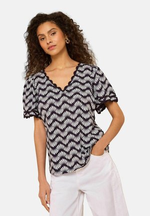 Kvinde med krøllet hår iført bluse med sort-hvidt bølgemønster og hvide bukser, en hånd i lommen, foran en enkel lys baggrund.