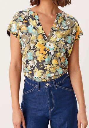 Nainen pukeutuneena lyhythihaiseen kukkakuosiseen puseroon, jossa on keltaisia, oransseja, sinivihreitä ja harmaita sävyjä, ja pusero on sullottu tummaan, korkeavyötäröiseen farkkuun.