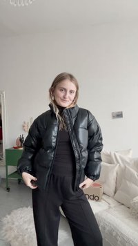 Czarna kurtka puchowa o błyszczącej fakturze, z wysokim kołnierzem i zapięciem na zamek. Połączona z czarną bluzką i luźnymi spodniami, stojąc w jasnym pomieszczeniu.