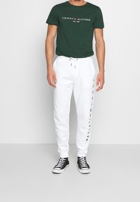 Man som bär en mörkgrön Tommy Hilfiger t-shirt och vita sweatpants med logotyp på benet, ihop med svarta högsula sneakers.